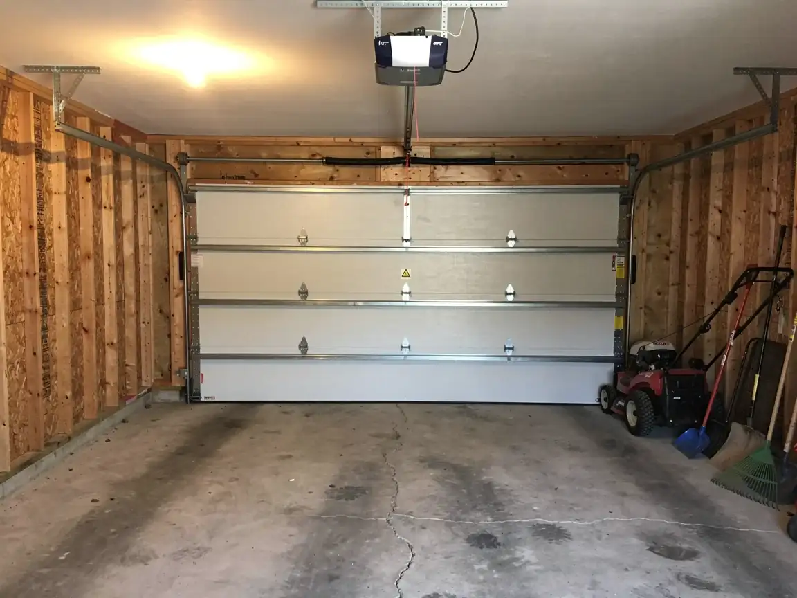 A white garage door is closed inside an unfinished garage with a lawnmower.